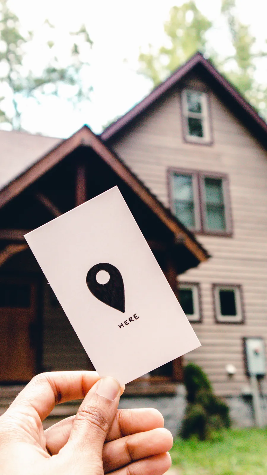 A hand holding a card with a location pin in front of a residential house.