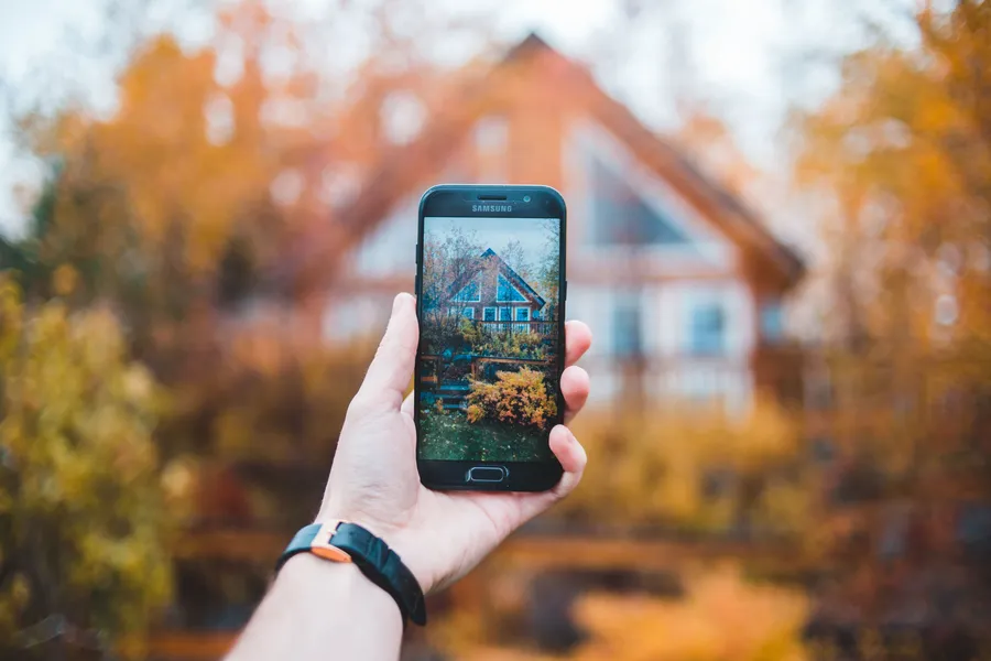 A hand holding a phone photographing a house, as if for a property listing.